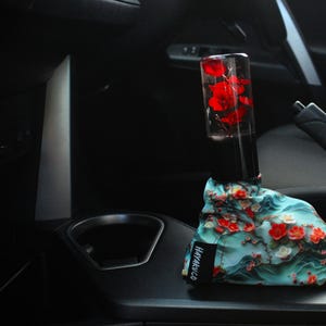 Könnte beinhalten: Ein blauer und weißer, floral gemusterter Schaltknauf für ein Auto mit rotem Blumendesign. Der Knauf ist an einem durchsichtigen Glaszylinder mit roten Blumen befestigt. Der Text "HAYABUSA" ist auf den Knauf gedruckt.