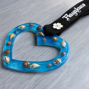 May include: A blue heart-shaped resin ornament with seashells embedded in it. The ornament has a black strap with the word "Haugljcuo" printed on it.
