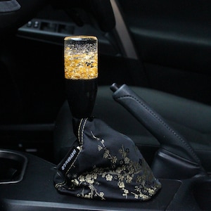 May include: A custom car shift knob with a clear resin top filled with gold flakes. The black base is paired with a black floral shift boot. The interior of a car is visible in the background.