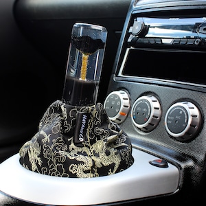 Könnte beinhalten: Ein individueller Autoschaltknauf mit einem transparenten, zylindrischen Design mit schwarzen und goldenen Akzenten. Die Schalthebelmanschette ist schwarz mit einem goldenen Drachenmuster. Installiert in einem Autoinnenraum.