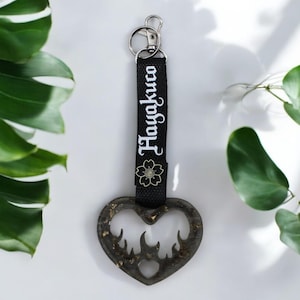 May include: Black heart-shaped keychain with a gold flame design and a black strap with the text "Hayakuko" in white.