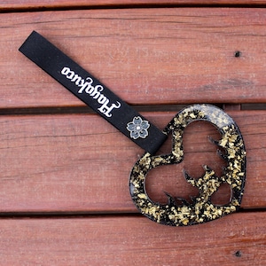 Könnte beinhalten: Schwarzer herzförmiger Schlüsselanhänger aus Harz mit goldenem Glitzer, der in das Harz eingebettet ist. Der Schlüsselanhänger hat ein schwarzes Band mit einem kleinen schwarzen Blumenanhänger und dem Text "Handcrafted" auf dem Band.