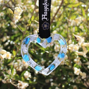 May include: A clear heart-shaped ornament with blue and white accents. The ornament is hanging from a black ribbon with the word "Hoyakuko" printed on it.
