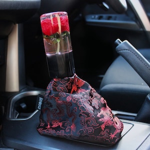 Op de afbeelding: Een zwarte versnellingspookknop met een rode roos ingekapseld in helder hars. De knop is bedekt met een zwarte stof met een rood drakenpatroon. De tekst "HAKKO" is zichtbaar aan de zijkant van de knop.