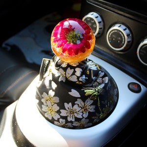 May include: A car gear shift knob featuring a resin flower design. The flower is pink and yellow and is encased in clear resin. The knob is covered in black fabric with a white floral pattern.