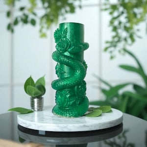 May include: A vibrant green decorative candle holder with a snake and floral design. The cylindrical holder is adorned with a detailed serpent coiled around it, with roses and leaves. The holder is displayed on a white marble-like surface.