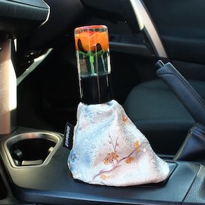 Puede incluir: Un pomo de palanca de cambios de coche con una parte superior de resina transparente que encierra una rosa naranja. La base negra se combina con una funda de palanca de cambios plateada con estampado floral. La funda de la palanca de cambios tiene una etiqueta que dice "HAYAKUCO".