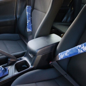 Puede incluir: El interior de un coche con dos asientos de cuero negro y una consola central. El asiento del pasajero delantero tiene una funda para el cinturón de seguridad con estampado floral azul y blanco. El asiento del conductor tiene una funda para el cinturón de seguridad azul.