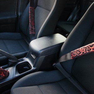 May include: Two black car seatbelt covers with a red and black floral pattern. The covers are installed in a black car interior.