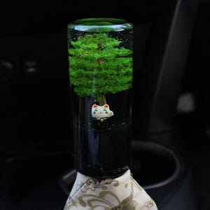 May include: A distinctive gear stick knob with a miniature green tree inside clear resin. A small, white cat figurine with red details is at the tree's base. The knob is mounted on patterned fabric.