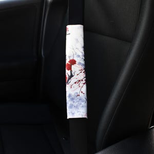 May include: A white car seatbelt cover with a floral pattern featuring red flowers and blue leaves. The cover is installed on a black car seat.