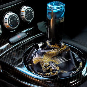 May include: A custom car interior with a shift knob featuring a blue and black swirl design. The shift boot is black with a gold and white dragon graphic. The console has carbon fibre accents and climate control dials.