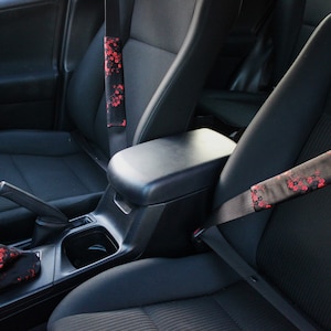 Könnte beinhalten: Schwarzer Auto-Innenraum mit Sicherheitsgurten und einem Schalthebel, die mit einem roten Blumenmuster verziert sind. Die Sicherheitsgurte haben passende Bezüge. Der Schaltknauf ist ebenfalls rot und schwarz.