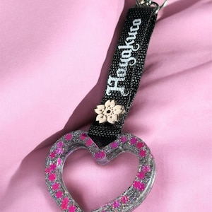 Puede incluir: Un llavero en forma de corazón con un marco de resina transparente relleno de purpurina plateada y pequeñas estrellas rosas. Incluye una correa de tela negra con el texto blanco Hayakuto y un pequeño dije de flor de cerezo.