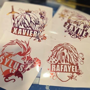 Puede incluir: Cuatro pegatinas blancas con diseños de lámina roja y dorada. Cada una presenta una cabeza estilizada con un nombre: Xavier, Zayne, Sylus y Rafayel. Los diseños incluyen elementos florales y decorativos.