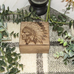 May include: A small, square wooden box with a detailed engraving of a Native American chief in profile on the lid. The box is a light brown color and sits on a patterned fabric, surrounded by green foliage.