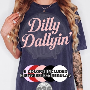 May include: Navy blue t-shirt with the text "Dilly Dallyin'" in pink script. The shirt includes a graphic with the text "5 COLORS INCLUDED DISTRESSED + REGULAR" and the shop logo. The shirt is a relaxed fit.