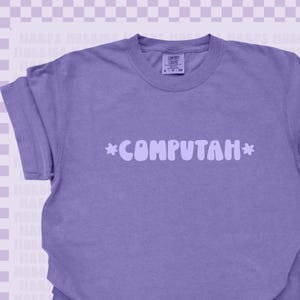 May include: A purple short-sleeve t-shirt featuring the text *COMPUTAH* in a light purple, rounded, bubbly font across the chest. The shirt has a crew neckline and a Comfort Colors brand label at the inner neck, indicating a size small.