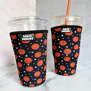 May include: Two clear plastic cups with orange straws, each encased in a black neoprene sleeve featuring a repeating pattern of orange basketballs and small white stars. A black rectangular tag with the white text Harps Hobbies is attached.