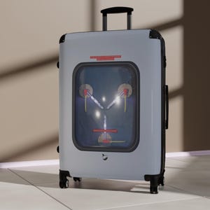 May include: A light gray rolling suitcase with black accents and wheels. The suitcase features a rectangular window with a futuristic design, including red and yellow details. The top handle is black.