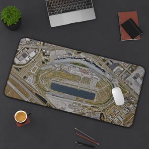 May include: A rectangular mouse mat showing an aerial view of a race track with a lake. The mat is on a dark surface with a laptop, mouse, pencils, a notebook, and a cup of coffee.