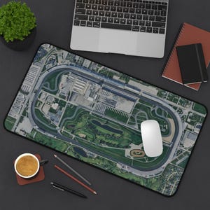 May include: A rectangular desk mat featuring an aerial view of a race track, with a black border. A white computer mouse sits on the mat. A laptop, notebooks, pencils, and a cup of coffee are also visible.