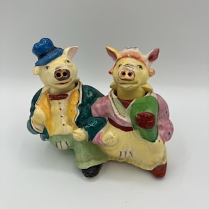 Puede incluir: Un par de figuras de cerdos de cerámica vintage. Un cerdo lleva un sombrero azul, una chaqueta verde y un chaleco amarillo. El otro cerdo lleva un vestido rosa y rojo y sostiene un objeto verde. Ambos tienen piel amarilla y están sonriendo.