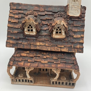 Może przedstawiać: Ręcznie wykonany ceramiczny domek z brązowym dachem z gontu, oknami w lukarnach i kominem. Dom posiada werandę z dekoracyjnymi podporami i szczegółową, teksturowaną powierzchnią. Paleta kolorów obejmuje odcienie brązu i beżu.
