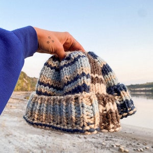 May include: A hand holds a blue, brown and white striped knitted beanie hat. The hat is made of thick yarn and has a slouchy fit.