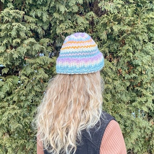 May include: A colorful striped knitted beanie hat in shades of blue, purple, white, and orange. The hat is worn by a person with long blonde hair.