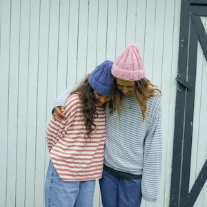 Peut inclure: Deux personnes portant des bonnets en tricot et des chemises à rayures. Un bonnet est bleu, l'autre rose. Les chemises ont des rayures horizontales rouges et blanches, et bleues et blanches. Elles s'embrassent devant un mur en bois blanc.