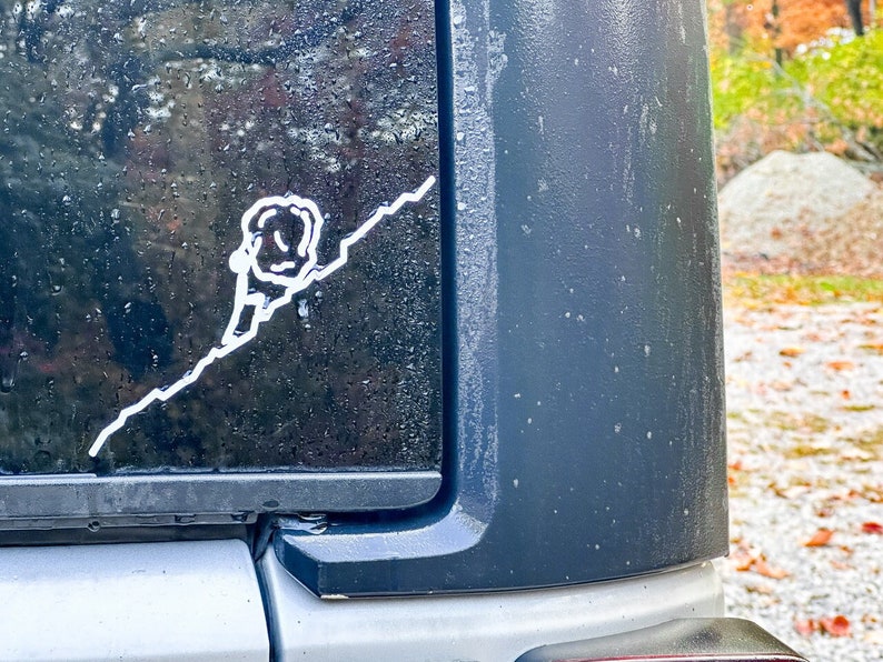 May include: White vinyl sticker of a person climbing a mountain, applied to a black vehicle surface. The sticker depicts a stylized climber with a rope ascending a diagonal line, suggesting adventure and outdoor activities.