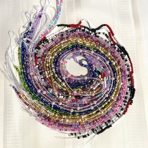 May include: A collection of colorful beaded waist beads, arranged in a spiral pattern. The beads are made of various materials, including glass, metal, and plastic. The beads are strung on a thin cord and are tied together at the ends. The beads are a variety of colors, including red, pink, purple, blue, green, yellow, and white.