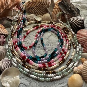 May include: A set of colourful beaded waist beads, featuring shades of white, pink, red, teal, black, and brown. The beads are arranged in a circular pattern and are displayed on a white background with seashells.