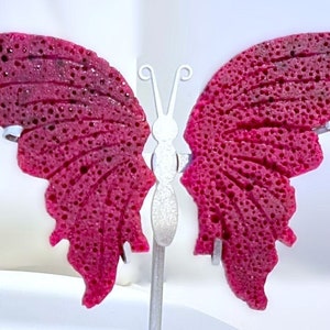 May include: A pink butterfly-shaped decorative object with a textured surface. The butterfly is mounted on a silver stand.
