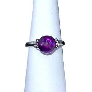 May include: A silver ring featuring a large, round, deep purple gemstone. The gemstone has a marbled appearance with lighter purple and white inclusions. The ring's band has decorative accents on either side of the gemstone.
