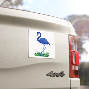 May include: A square white magnet featuring a blue flamingo standing in green grass. The flamingo has a curved neck and is set against a white background. The magnet is attached to a vehicle.