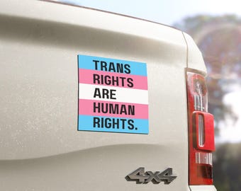 May include: A white truck with a blue, pink, and white transgender pride flag sticker on the back. The sticker says "Trans Rights Are Human Rights."