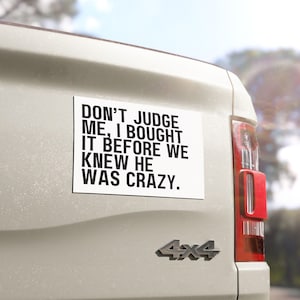 May include: A white rectangular bumper sticker with black text that reads "DON'T JUDGE ME, I BOUGHT IT BEFORE WE KNEW HE WAS CRAZY." The sticker is affixed to the back of a light-colored vehicle, near the taillight and a 4x4 emblem.