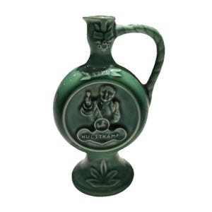 Może przedstawiać: Zielony ceramiczny dzban z uchwytem i okrągłym korpusem. Dzbanek posiada relief przedstawiający osobę oraz napis "HULSTKAMP". Widoczny jest również rok "1775". Dzbanek posiada dekoracyjną podstawę i wylewkę.