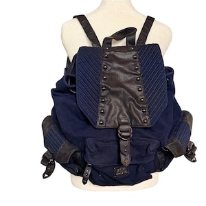 Op de afbeelding: Een marineblauwe rugzak met een klepsluiting en meerdere vakken. De klep en accenten zijn donkerbruin leer met metalen stud details. De rugzak heeft verstelbare bandjes en een trekkoordsluiting.