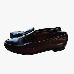 May include: A pair of dark burgundy leather penny loafers. The shoes have a glossy finish and a classic design, featuring a strap across the vamp with a slit for a penny. The soles are black.
