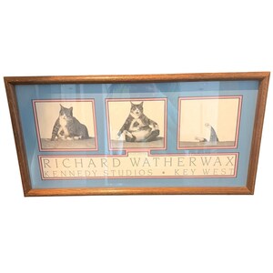 Könnte beinhalten: Ein rechteckiger Holzrahmen mit drei schwarz-weißen Katzenfotos auf blauem Hintergrund mit rosa Rändern. Der Text lautet: RICHARD WATHERWAX KENNEDY STUDIOS • KEY WEST. Die Bilder zeigen eine sitzende Katze, eine aufrecht sitzende Katze und ein Katzenbein.