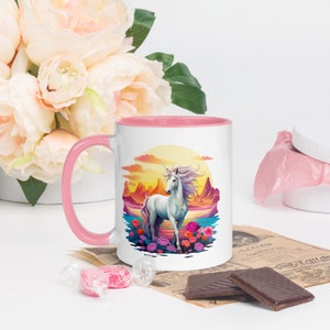 May include: A white ceramic mug with a pink rim and handle. The mug features a colorful illustration of a white unicorn standing in a field of flowers against a sunset backdrop.
