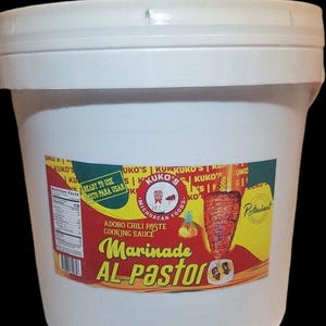 May include: A white plastic bucket with a red and yellow label. The label says "Kuko's Michoacan Foods" and "Marinade Al Pastor". The label also has a picture of a taco and a pineapple.