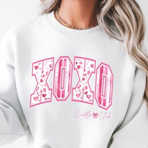 May include: White sweatshirt with the letters "XOXO" in pink, filled with heart patterns. The sweatshirt also features the text "Cuddle Club" in a script font. The design is centered on the front of the garment.