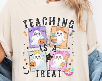 May include: Cream-colored t-shirt featuring a Halloween-themed graphic. The design includes four framed images of cartoon ghosts, pumpkins, and the text "TEACHING IS A TREAT" in black. The shirt is a casual, everyday wear item.
