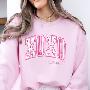May include: A light pink sweatshirt with the word "XOXO" in a pink and white design filled with hearts. The text "Cuddle Club" is below the main text. The sweatshirt is a soft, comfortable material.
