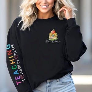 Può includere: Felpa nera con un testo arcobaleno "Teaching, A Work of Heart" sulla manica sinistra e una pila di libri con una mela rossa in cima e un fiocco giallo sui libri. Il testo "Miss. Roberts" è sotto i libri.