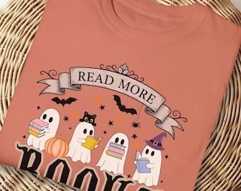 May include: Coral-colored t-shirt featuring a Halloween-themed graphic. The design includes cartoon ghosts reading books, a pumpkin, bats, stars, and the text "READ MORE BOOKS". The shirt has a round neckline and short sleeves.